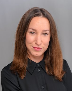 A woman with long brown hair is wearing a black shirt and looking directly at the camera. The background is gray.