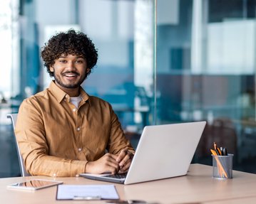 A young man with curly hair sits at a desk, smiling as he works on a laptop. Writing utensils are visible.