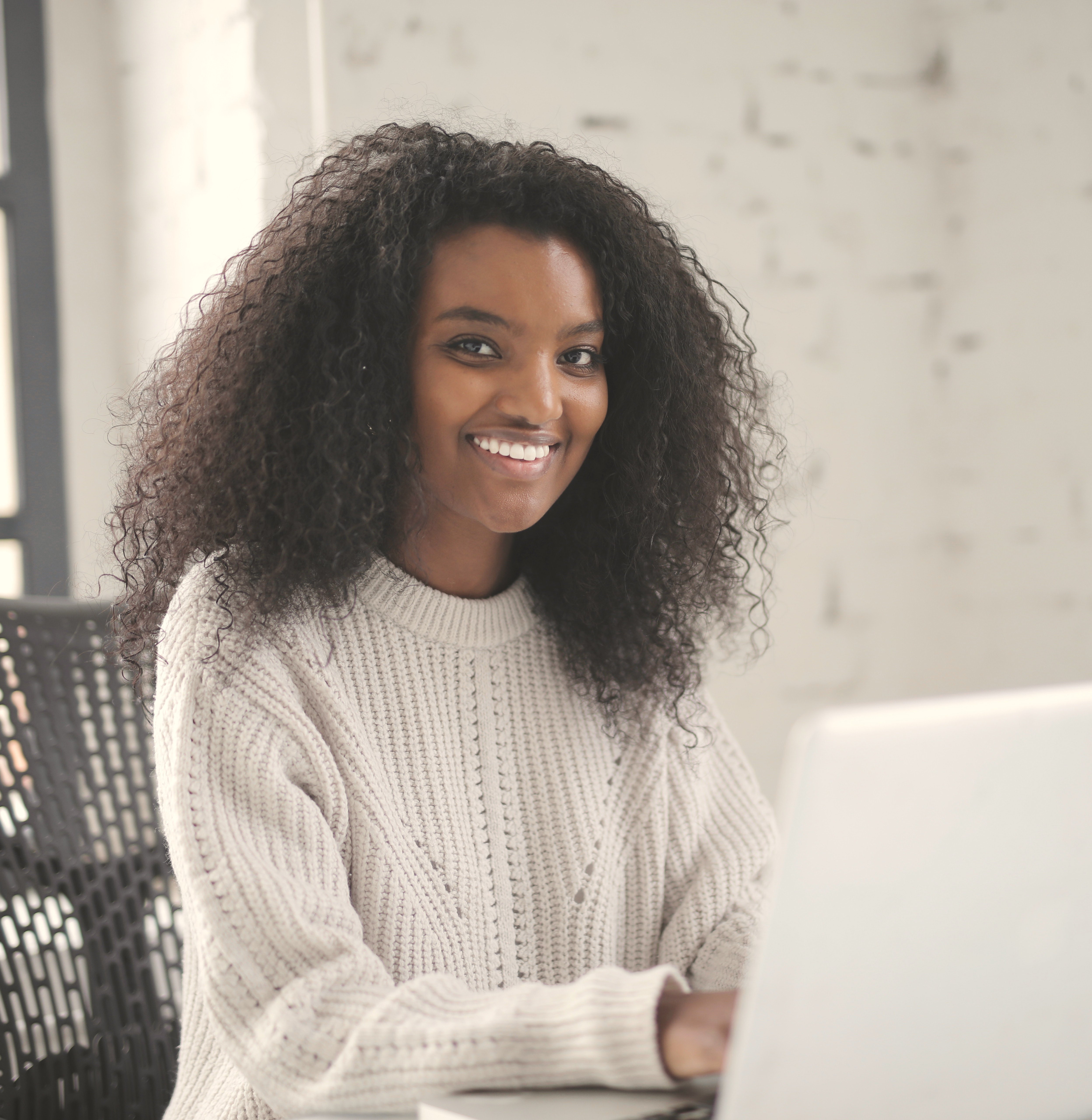Eine Person mit lockigem, schwarzem Haar sitzt an einem Schreibtisch und arbeitet an einem Laptop. Sie trägt einen hellen, gestrickten Pullover und lächelt in die Kamera. Der Hintergrund zeigt eine helle, minimalistische Umgebung.