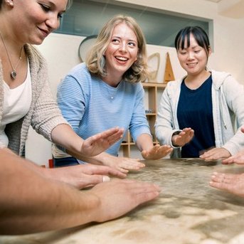 Fünf Personen stehen um einen Tisch und legen ihre Hände darauf. Sie scheinen an einer Aktivität oder Übung beteiligt zu sein.