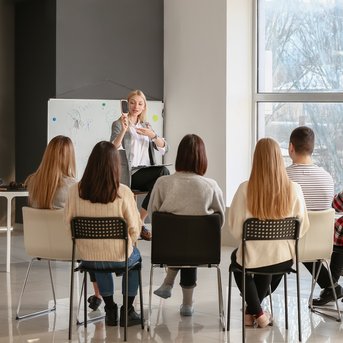 Eine Gruppe von Menschen sitzt in einem Seminarraum und hört einer Referentin zu, die an einer Tafel spricht.