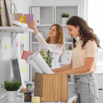 Two women are organizing a desk. One is putting folders on the shelf while the other is taking plans out of a box.