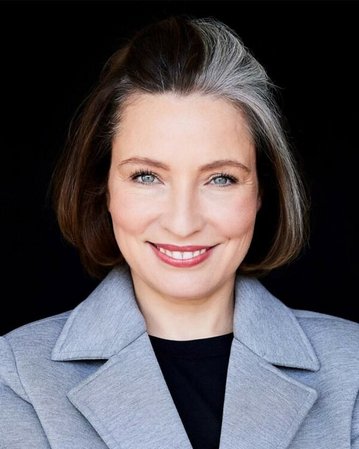 Eine Frau mit kurzen, braunen Haaren und grauen Strähnen lächelt in die Kamera, trägt einen grauen Blazer und hat einen neutralen Hintergrund.