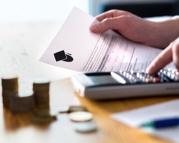 A hand holds a document with a graduation cap symbol, while the other hand operates a calculator. Coins are lying on the table.