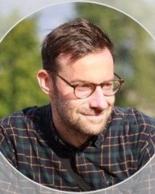 Ein Mann mit Brille und kariertem Hemd, der nachdenklich schaut. Unscharfer Hintergrund mit grünen Bäumen.