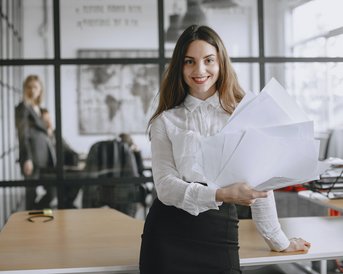 Eine Frau in einem Büro hält mehrere Blätter Papier in der Hand und lächelt. Im Hintergrund steht eine weitere Person, die an einem Schreibtisch arbeitet. Der Raum hat große Fenster und moderne Büromöbel.