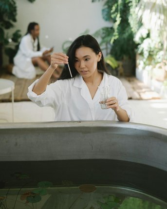Eine Frau in einem weißen Kittel hält eine Pipette über ein Wasserbecken mit Pflanzen. Im Hintergrund sitzt eine andere Person in einem weißen Outfit. Die Umgebung ist grün und pflanzenreich.