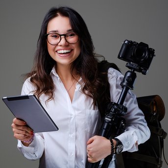 Eine lächelnde Frau mit Brille hält ein Tablet in der Hand und steht neben einem Stativ mit einer Kamera. Sie trägt einen Rucksack.