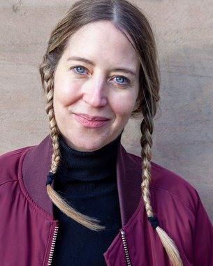 A woman with long, braided pigtails is wearing a burgundy bomber jacket and a black turtleneck sweater. She is looking directly at the camera and has blue eyes. The background is neutral and unobtrusive.
