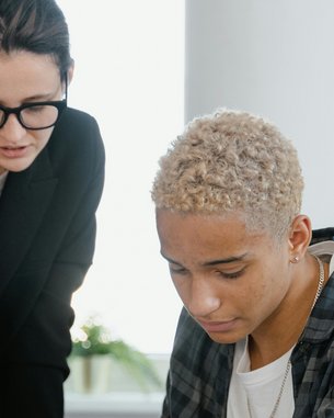 Eine Frau mit Brille erklärt einem Mann mit blonden Locken etwas, während er auf Dokumente auf einem Tisch schaut. Auf dem Tisch liegen mehrere Papiere und Schreibutensilien. Ein Glas Wasser steht ebenfalls auf dem Tisch.