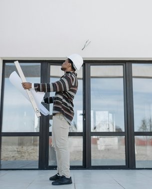 Ein Bauarbeiter mit einem Helm hält Baupläne in der Hand und steht in einem leeren Raum mit großen Fenstern.