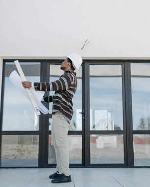Ein Bauarbeiter mit einem Helm hält Baupläne in der Hand und steht in einem leeren Raum mit großen Fenstern.