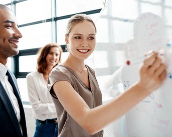 Drei Personen in einem modernen Büro, eine Frau schreibt an eine Glaswand, während zwei andere sie anlächeln. Sie scheinen an einem kreativen Projekt zu arbeiten.