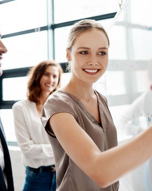 Drei Personen in einem modernen Büro, eine Frau schreibt an eine Glaswand, während zwei andere sie anlächeln. Sie scheinen an einem kreativen Projekt zu arbeiten.