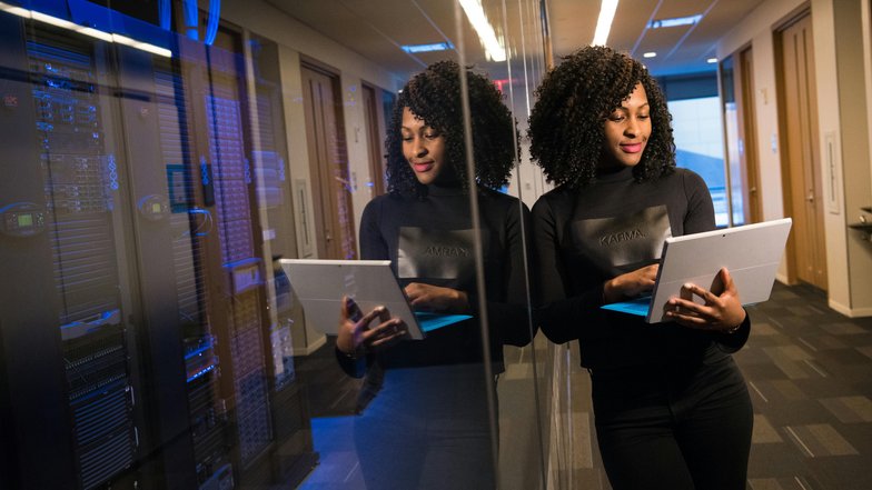 Eine Frau mit lockigem Haar steht in einem modernen Büro und betrachtet ein Tablet. Sie trägt ein schwarzes Oberteil und steht vor einer reflektierenden Wand. Im Hintergrund sind Server zu sehen.