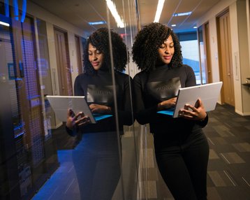 Kategorie Data Science studieren Studium SRH University Eine Frau mit lockigem Haar steht in einem modernen Büro und betrachtet ein Tablet. Sie trägt ein schwarzes Oberteil und steht vor einer reflektierenden Wand. Im Hintergrund sind Server zu sehen.