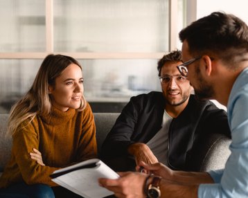 Two people are sitting on a sofa and having a conversation. A woman with long hair is wearing a brown sweater, while a man in a black top is smiling. A third man is holding a notebook and talking to them.
