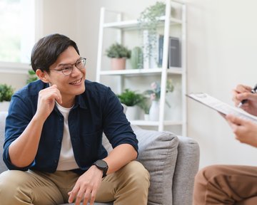 Ein junger Mann mit Brille sitzt auf einem Sofa und lächelt, während er einem Gesprächspartner gegenübersitzt, der Notizen macht.
