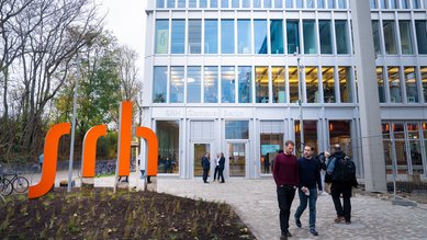 Zwei Personen gehen an einem modernen Gebäude mit großen Fenstern vorbei, vor dem ein orangefarbenes Schild mit der Aufschrift "srh" steht. Bäume und Pflanzen sind im Vordergrund sichtbar.