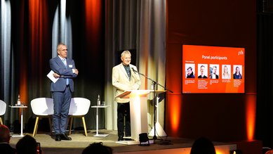 Ein Mann steht am Rednerpult und spricht, während ein anderer Mann daneben steht. Im Hintergrund sind Stühle und ein Bildschirm mit Informationen zu den Panelteilnehmern sichtbar.