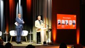 Ein Mann steht am Rednerpult und spricht, während ein anderer Mann daneben steht. Im Hintergrund sind Stühle und ein Bildschirm mit Informationen zu den Panelteilnehmern sichtbar.