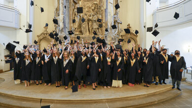 A group of graduates in academic attire throwing their hats in the air, set against an ornate backdrop in a hall.