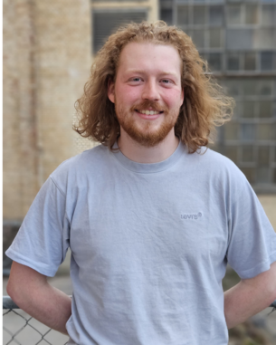 Henning Tholen steht lächelnd mit verschränkten Armen in einem hellen T-Shirt vor einer Wand mit Fenstern.