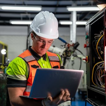Ein Arbeiter in Sicherheitskleidung und Helm überprüft ein Laptop in einer industriellen Umgebung mit elektrischen Geräten.