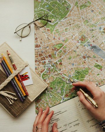 A person is holding a pen and pointing at a city map that is laid out on a table. Next to the map, there is a notebook and some colorful pens. A pair of glasses is also on the table.
