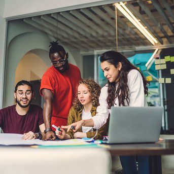 Four people are working together at a table, surrounded by notes and a laptop. They are discussing and sketching on a large sheet of paper as they exchange ideas and collaborate on a project.