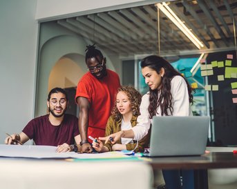 Four people are working together at a table, surrounded by notes and a laptop. They are discussing and sketching on a large sheet of paper as they exchange ideas and collaborate on a project.