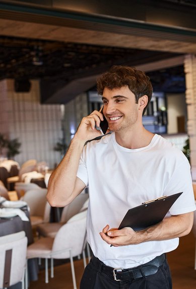 Ein junger Mann spricht am Telefon und hält einen Notizblock in einem eleganten Restaurant mit gedeckten Tischen.