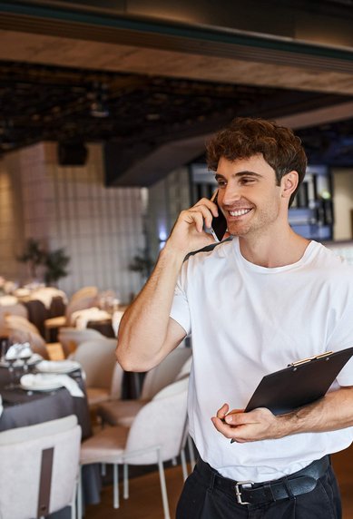 Ein junger Mann spricht am Telefon und hält einen Notizblock in einem eleganten Restaurant mit gedeckten Tischen.