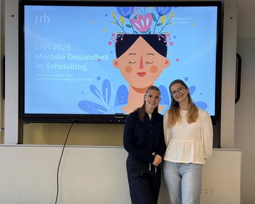 Zwei Frauen stehen vor einem Bildschirm, der Informationen über mentale Gesundheit im Schulalltag zeigt.