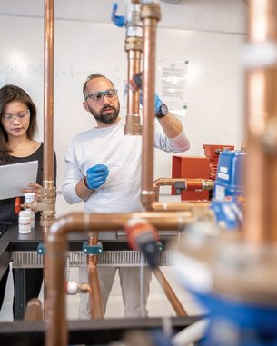 Eine Frau mit Brille hält ein Blatt Papier, während ein Mann mit Bart und Schutzbrille an einer Kupferrohrinstallation arbeitet. Im Hintergrund sind technische Geräte und eine Tafel sichtbar.