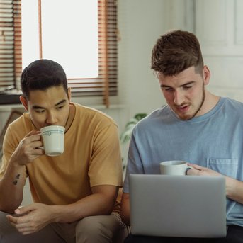 Zwei Männer sitzen auf einem Bett, trinken Kaffee und schauen auf einen Laptop. Helle, gemütliche Umgebung mit Fensterlicht.