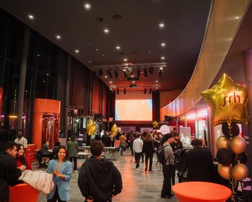 Eine belebte Veranstaltungshalle mit Gästen, orangefarbenen Tischen und goldenen Sternballons. Im Hintergrund ist eine Präsentation auf einer Leinwand zu sehen.