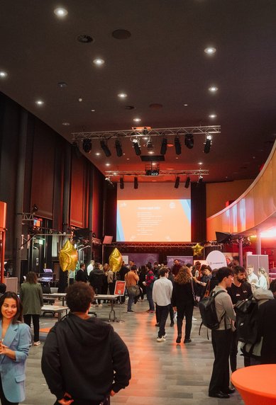 A bustling event hall filled with guests, orange tables, and golden star balloons. In the background, a presentation is displayed on a screen.