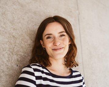 A young woman with curly brown hair smiles warmly. She is wearing a striped top and stands in front of a gray wall. Her eyes are bright and expressive, giving her an open appearance.