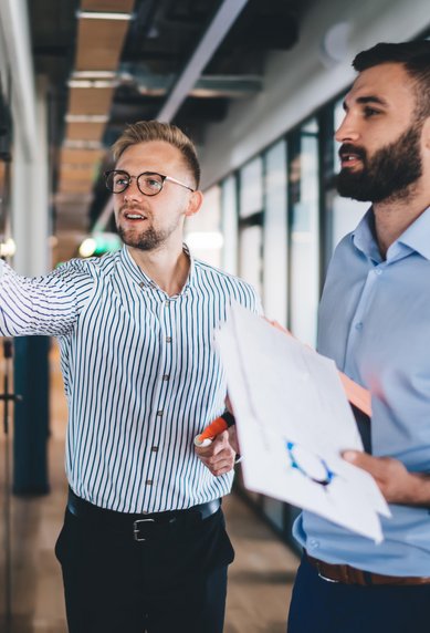 Zwei Männer in einem modernen Büro, einer schreibt an einer Glaswand, der andere hält Unterlagen und beobachtet.