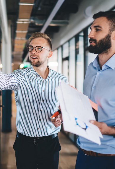 Two men in a modern office, one is writing on a glass wall while the other holds documents and observes.