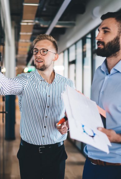 Zwei Männer in einem modernen Büro, einer schreibt an einer Glaswand, der andere hält Unterlagen und beobachtet.