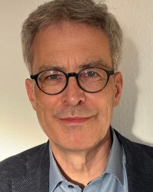 Ein Mann mit Brille und grauen Haaren lächelt in die Kamera, trägt ein blaues Hemd und einen dunkelgrauen Blazer.