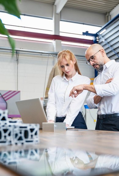 Two people in a modern office are discussing information on a laptop while standing at a table with technical components.