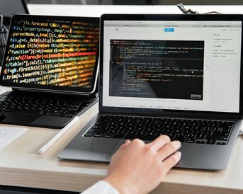 Ein Laptop mit Code auf dem Bildschirm und ein Tablet mit bunten Programmierzeilen im Hintergrund. Eine Hand tippt auf der Laptop-Tastatur.