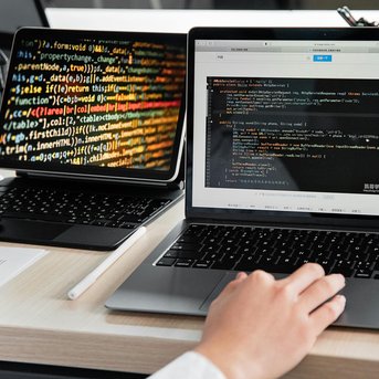 Ein Laptop mit Code auf dem Bildschirm und ein Tablet mit bunten Programmierzeilen im Hintergrund. Eine Hand tippt auf der Laptop-Tastatur.