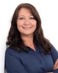 Eine Frau mit langen, dunklen Haaren und einem blauen Oberteil lächelt freundlich und steht vor einem weißen Hintergrund.