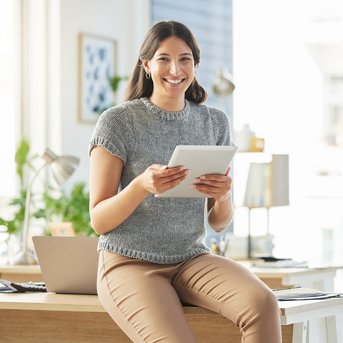 Eine lächelnde Frau sitzt auf einem Tisch und hält ein Tablet in der Hand. Sie trägt ein graues Oberteil und hat lange, lockige Haare. Im Hintergrund sind Büromöbel und Pflanzen zu sehen.