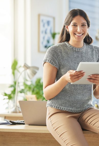 Eine lächelnde Frau sitzt auf einem Tisch und hält ein Tablet in der Hand. Sie trägt ein graues Oberteil und hat lange, lockige Haare. Im Hintergrund sind Büromöbel und Pflanzen zu sehen.