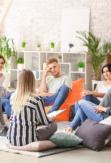Eine Gruppe von sieben Personen sitzt in einem modernen Raum auf Sitzsäcken und diskutiert miteinander. Pflanzen und Regale sind im Hintergrund sichtbar.